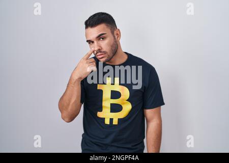 Jeune homme hispanique portant le t-shirt bitcoin pointant vers l'œil vous regardant le geste, l'expression suspecte Banque D'Images