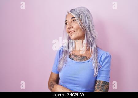 Femme d'âge moyen avec tatouages debout sur fond rose regardant loin à côté avec le sourire sur le visage, expression naturelle. rire confiant. Banque D'Images
