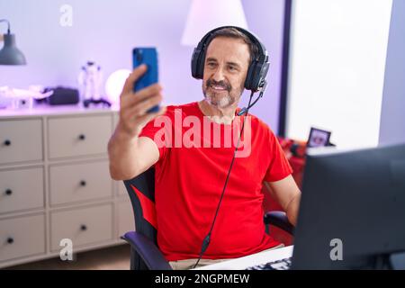 Homme d'âge moyen, streamer souriant et confiant, faites le selfie par smartphone dans la salle de jeux Banque D'Images