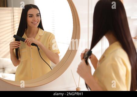 Jeune femme redressant les cheveux près du miroir à la maison. Routine du matin Banque D'Images