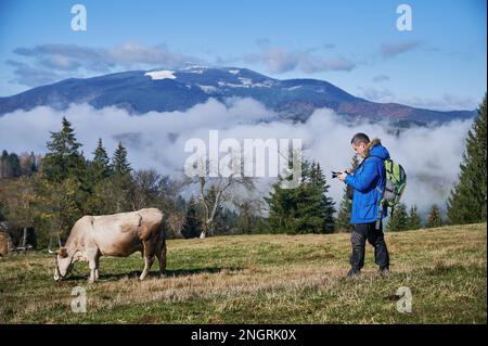 Photographe masculin prenant la photo d'un animal de ferme sur un pré alpine herbacé avec des montagnes brumeuses en arrière-plan Banque D'Images