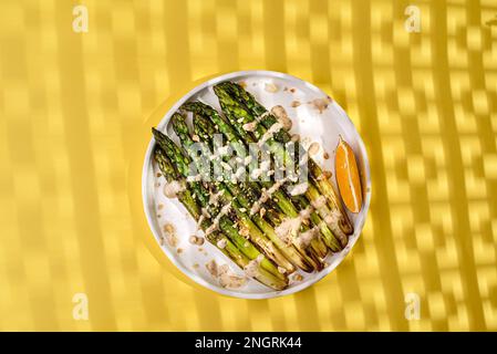 asperges cuites dans une assiette. asperges frites à l'huile d'olive, assaisonnées de sauce et saupoudrées de noix, à côté d'une tranche de citron. Sur un fond jaune Banque D'Images