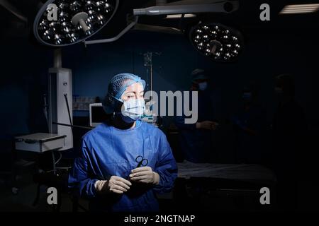 Portrait éclairé d'une infirmière de salle d'opération portant un EPI avec des ciseaux chirurgicaux dans les mains debout dans la salle d'opération avec l'équipe chirurgicale Banque D'Images