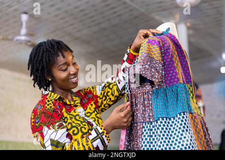 Souriant couturier ghanéen aux cheveux locs mesure une partie de sa robe africaine colorée dans son studio Banque D'Images