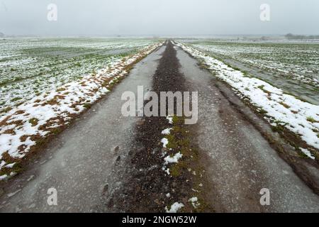 Route de terre glacée à travers les champs avec de la neige et de l'eau, vue sur un jour brumeux, Czulczyce, Pologne Banque D'Images