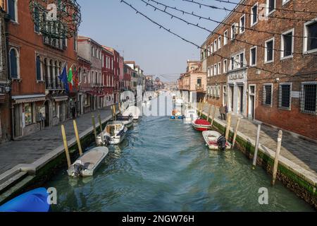Murano, Italie, vue de jour sur un canal étroit avec des bâtiments bas en couleur, des boutiques et des bateaux amarrés sur des quais en bois. Banque D'Images