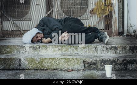 Un homme sans abri gelé dort sur du béton près du système de ventilation du bâtiment. Problème de la façon dont les sans-abri survivent au froid hivernal. Banque D'Images