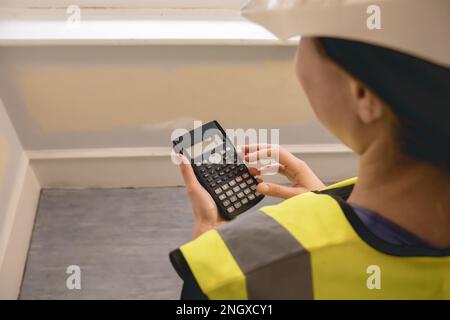Femme d'ingénieur civil méconnaissable calculant avec une calculatrice dans un chantier de construction, porte un gilet jaune haute visibilité, un epi, un casque, un casque Banque D'Images