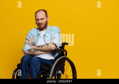 Un homme dans un fauteuil roulant problèmes avec le système musculo-squelettique regarde la caméra n'est pas satisfait, avec des tatouages sur ses bras assis sur un studio jaune Banque D'Images