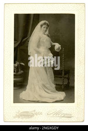 Original Edwardian Era studio portrait de carte de mariage d'une belle jeune mariée portant une longue robe blanche, avec un décolleté haut, et long voile, tenant un bouquet de fleurs. Les styles de manches et d'encolure datent de la photographie vers 1900-1902. Mariage édouardien. Mariée édouardienne. Du studio de G (George William) Morgan, 393 Union Street, Aberdeen, Écosse, Royaume-Uni Banque D'Images