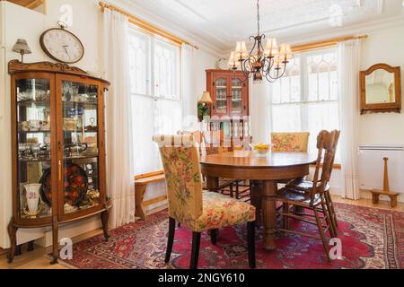 Table à manger ovale en chêne avec chaises à dossier, buffet d'angle en bois avec portes vitrées, tapis rouge cramoisi dans la salle à manger avec lustre ancien éclairé. Banque D'Images