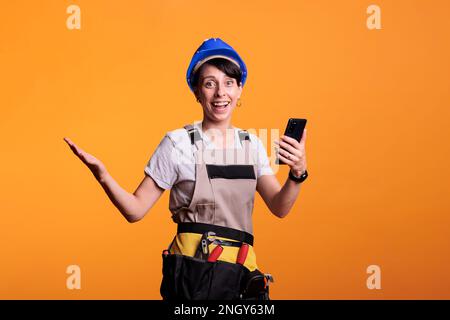 Un ouvrier du bâtiment souriant à l'application pour smartphone, surfant sur Internet et posant en studio. Être heureux et regarder le site Web de téléphone mobile, porter un casque et rénover des combinaisons. Banque D'Images