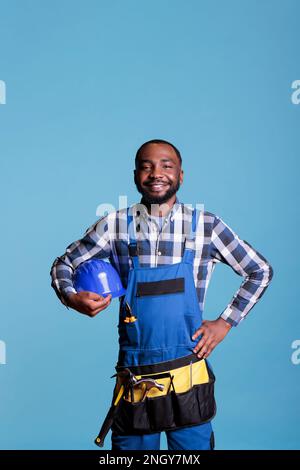 Travailleur de la construction afro-américain qui se réjouit du lieu de travail et qui tient un casque de sécurité. Homme de main souriant portant une combinaison et une ceinture à outils isolée sur fond bleu, prise de vue en studio. Banque D'Images