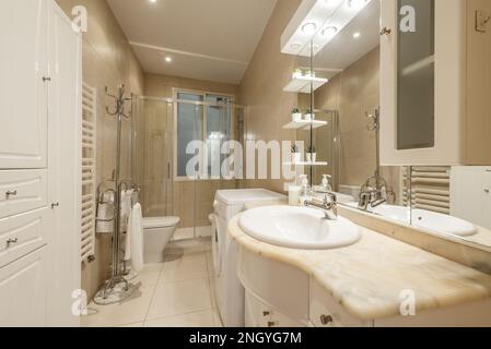 Salle de bains avec lavabo en marbre crème et armoire en bois avec miroir intégré avec lumières et étagères et lave-linge dans un coin Banque D'Images