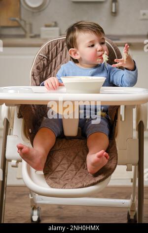 Un enfant drôle mange une pomme râpée avec sa bouche pleine tout en étant assis sur une chaise de cuisine. Bébé garçon affamé, il a de la nourriture dans sa bouche, de l'humour. Enfant âgé Banque D'Images