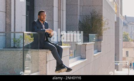 Homme afro-américain millénaire en costume d'affaires assis à l'extérieur près du bâtiment de bureau boire du thé chaud. Entrepreneur ethnique confiant Banque D'Images
