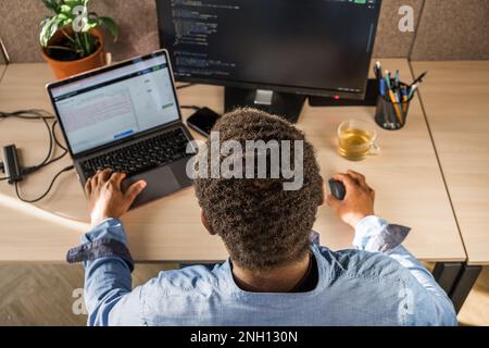 Black Man écrit des lignes de code sur un PC de bureau avec deux moniteurs et un ordinateur portable mis de côté dans un bureau élégant. Programmation de développeur masculin professionnel Banque D'Images