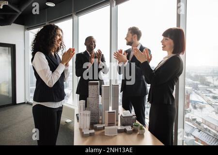 Bons résultats, félicitations et concept d'appréciation. Satisfait divers hommes d'affaires et femmes en tenue formelle, debout près de la table avec la maquette de bâtiment de gratte-ciel dans la salle de conférence et applaudissant Banque D'Images