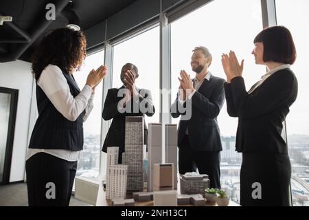 Bons résultats, félicitations et concept d'appréciation. Satisfait divers hommes d'affaires et femmes en tenue formelle, debout près de la table avec la maquette de bâtiment de gratte-ciel dans la salle de conférence et applaudissant Banque D'Images