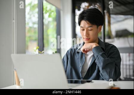 Jeune asiatique concentré regardant l'écran de tablette, pensivement pensant à son projet de marketing, travaillant dans le bureau. Banque D'Images