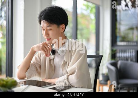 Jeune asiatique élégant étudiant ou indépendant dans des vêtements décontractés se concentrant sur son projet, en utilisant une tablette numérique, travaillant à distance au café Banque D'Images