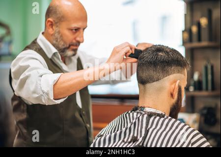 coiffeur mâle barbu avec peigne et ciseaux faisant une coupe de cheveux à l'homme dans le salon contemporain Banque D'Images