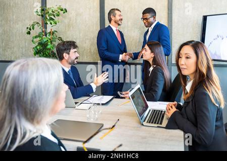 Un groupe de collègues multiethniques se réunit dans une salle de réunion, tandis qu'un homme noir se secoue avec un collègue et une femme asiatique assis à table avec un talki pour ordinateur portable Banque D'Images