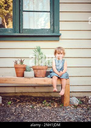 Le tout-petit est assis sur un banc avec des plantes en pot dans la cour de la ferme. Banque D'Images