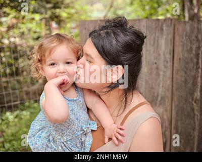 La mère embrasse la fille sur la joue, la fille regarde l'appareil photo Banque D'Images