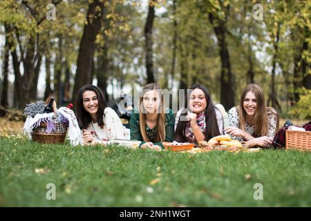 Groupe de quatre femmes ayant un pique-nique amusant dans le parc, en posant sur une couverture, en appréciant la nourriture et la nature. Banque D'Images