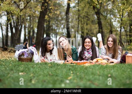 Groupe de quatre femmes ayant un pique-nique amusant dans le parc, en posant sur une couverture, en appréciant la nourriture et la nature. Banque D'Images