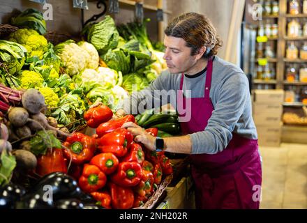 Un travailleur d'épicerie de sexe masculin organise des poivrons et d'autres légumes sur le comptoir et le présentoir Banque D'Images