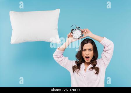 femme choquée en pyjama tenant un réveil vintage au-dessus de la tête près de lévitant oreiller sur bleu, image de stock Banque D'Images