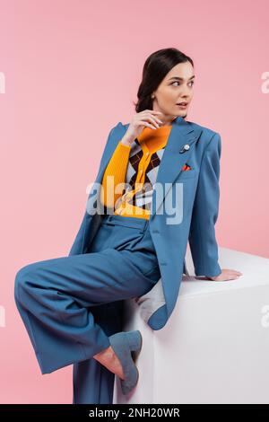 jolie femme en costume bleu posant près du cube blanc et regardant loin isolé sur rose, image de stock Banque D'Images