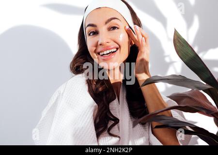 Une femme brune heureuse dans un peignoir blanc prend soin de sa peau et hydrate son visage avec une crème. Une jeune femme positive bénéficie d'un soin de beauté Banque D'Images