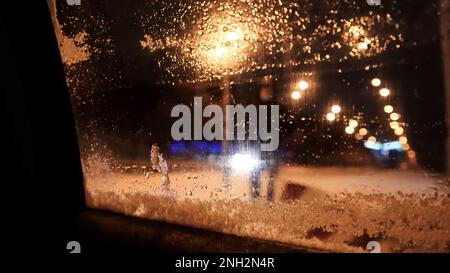 Vitre de voiture embuée. Fonte de neige sur une vitre de voiture. Phares de voiture, bokeh et reflets de couleur vive derrière le verre recouvert de neige Banque D'Images