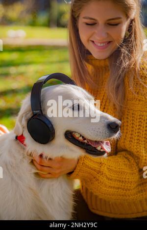 Belgrade, Serbie. 10 novembre 2022. Une jeune fille souriante et son adorable retriever d'or portant des écouteurs noirs dans un parc par temps ensoleillé. Banque D'Images