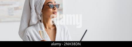 femme afro-américaine en robe blanche et lunettes de soleil élégantes tenant le champagne et la lime à ongles tout en regardant à la maison, bannière, image de stock Banque D'Images