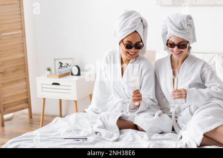 femmes multiethniques heureuses dans des peignoirs doux et des lunettes de soleil assis avec des verres à champagne sur le lit, image de stock Banque D'Images