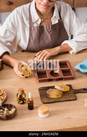 vue rognée de l'artisan afro-américain tenant du savon à base de plantes près des moules en silicone et des ingrédients naturels, image de stock Banque D'Images