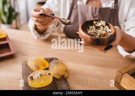 vue rognée de l'artisan afro-américain tenant un bol en bois et une cuillère avec des plantes séchées près de savon maison à base de plantes, image de stock Banque D'Images