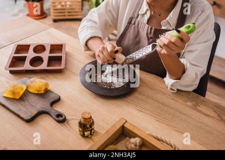 vue rognée de l'artisan afro-américain râper savon près de moule en silicone et huile essentielle sur table en bois, image de stock Banque D'Images