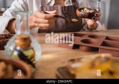 vue rognée de l'artisan afro-américain tenant un bol en bois et une cuillère avec des herbes séchées près de moule en silicone sur un premier plan flou, image de stock Banque D'Images