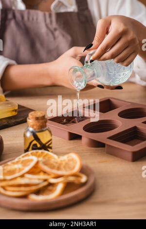 vue rognée de l'artisan afro-américain versant du savon liquide dans le moule en silicone près des tranches d'orange séchées et de l'huile essentielle, image de stock Banque D'Images