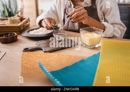 vue rognée de l'artisan afro-américain trempant bougie mèche dans la cire d'abeille fondue près de planche à découper et feuilles de cire, image de stock Banque D'Images