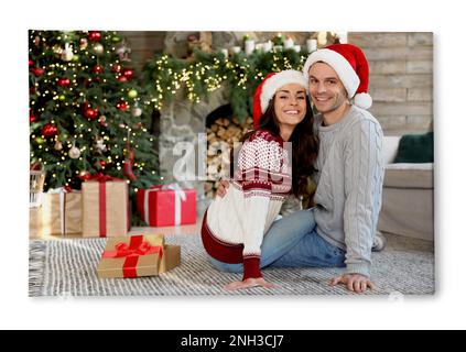 Photo imprimée sur toile, fond blanc. Joyeux jeune couple portant des chapeaux de père Noël dans le salon décoré pour Noël Banque D'Images