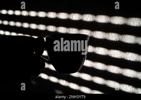 Silhouette d'une tasse à café avec diffusion de lumière à travers les volets de fenêtre sur le mur Banque D'Images