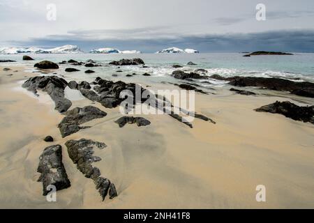 Paysage côtier avec des formations rocheuses entourées de vagues douces et de montagnes lointaines enneigées sous un ciel gris. Banque D'Images