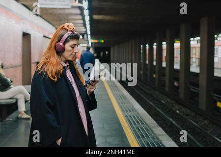 Une jeune femme ennuyeuse attend que le métro écoute de la musique dans les écouteurs de son smartphone Banque D'Images