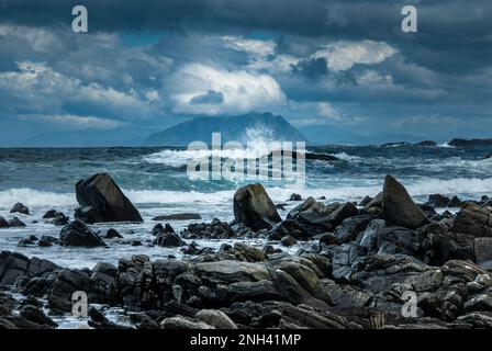 Les vagues s'écrasent énergiquement contre des formations rocheuses tandis qu'une île lointaine est visible sous un ciel nuageux. Banque D'Images